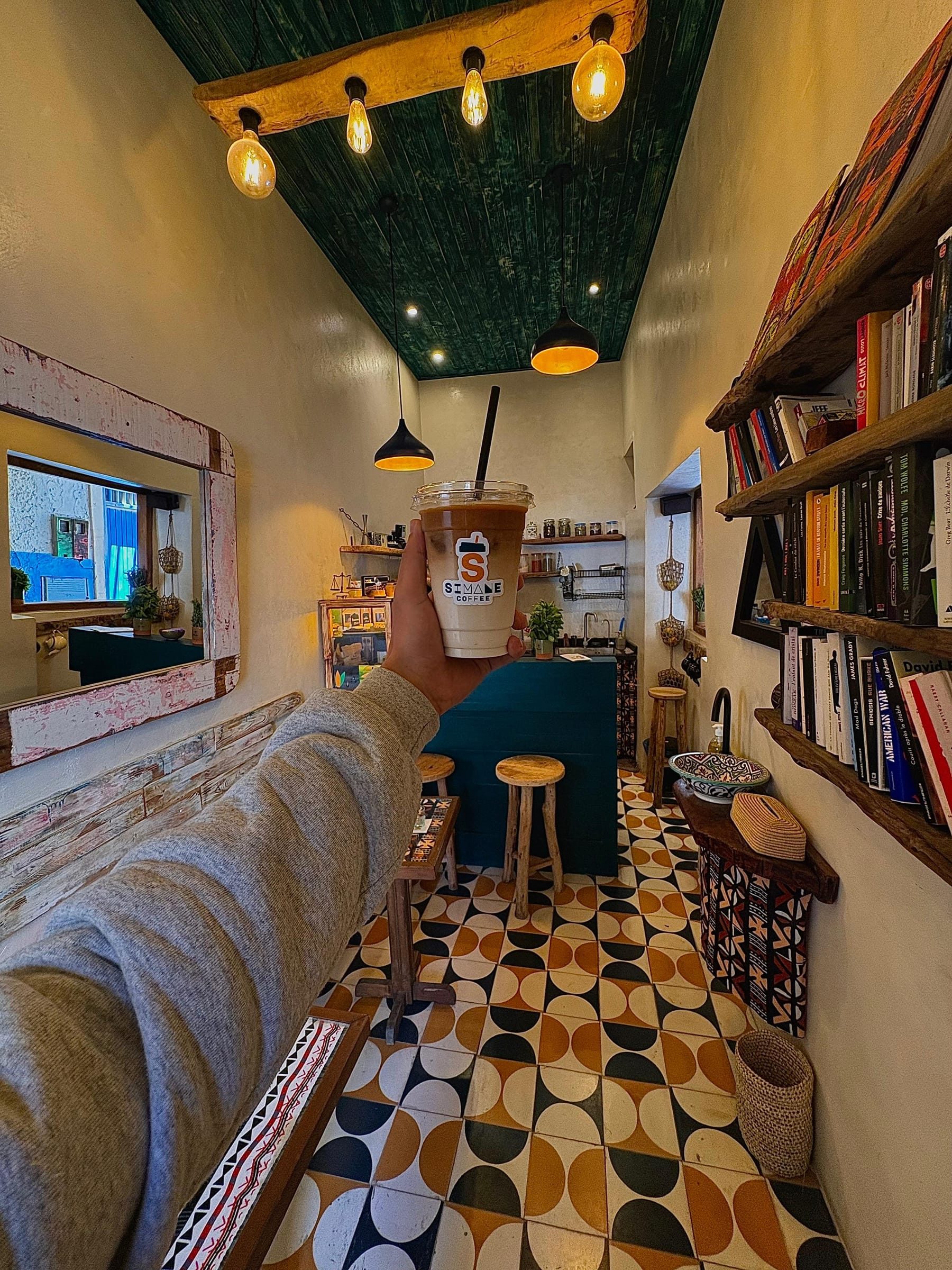 A hand holding an iced coffee inside Simane, the counter and bookshelves behind